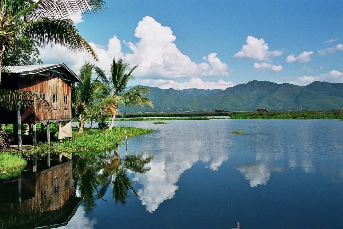 A wooden stilt house and palm trees line the calm waters of Inle Lake, with mountains reflected under a blue sky.