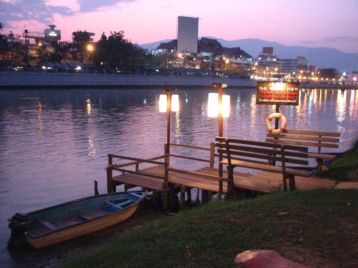 Twilight settles over the Ping River in Chiang Mai with a wooden pier, lanterns, and a small boat by the bank.