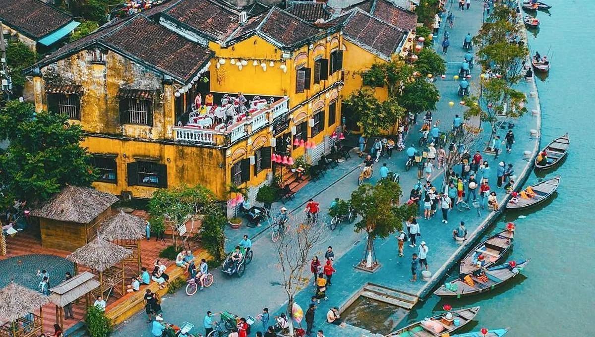 Aerial view of Hoi An's yellow ancient houses along a riverside street bustling with visitors and boats.
