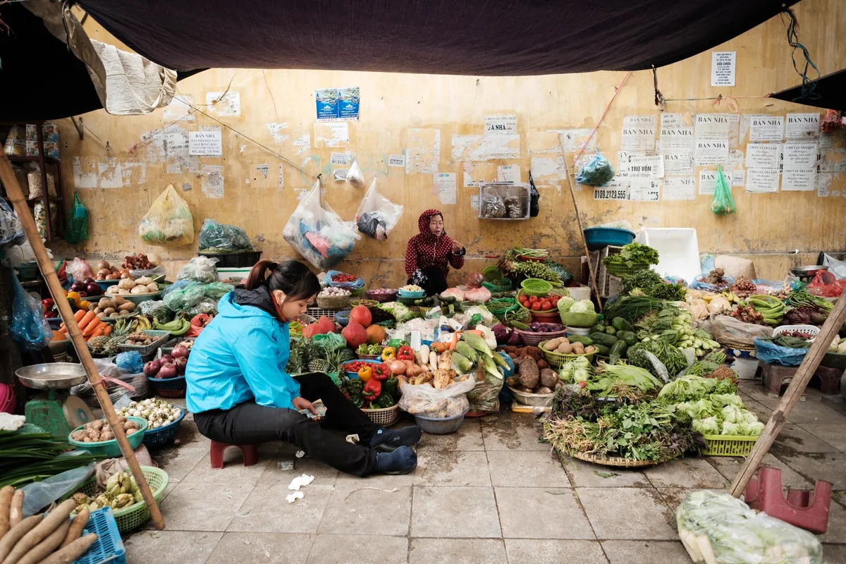 Vendors sit among piles of fresh vegetables at a covered market stall in Hanoi's Old Quarter.
