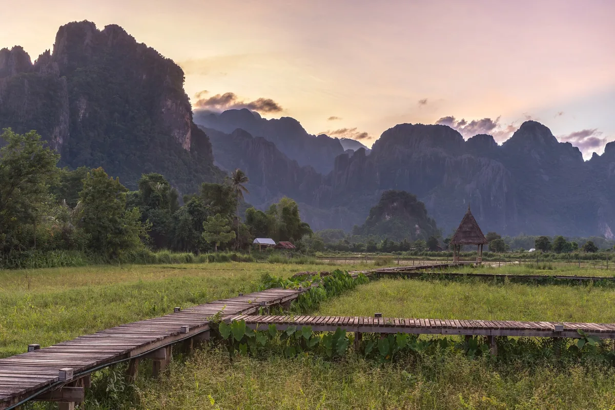 A wooden walkway winds through green rice fields toward a small hut beneath karst mountains at sunset in Vang Vieng.