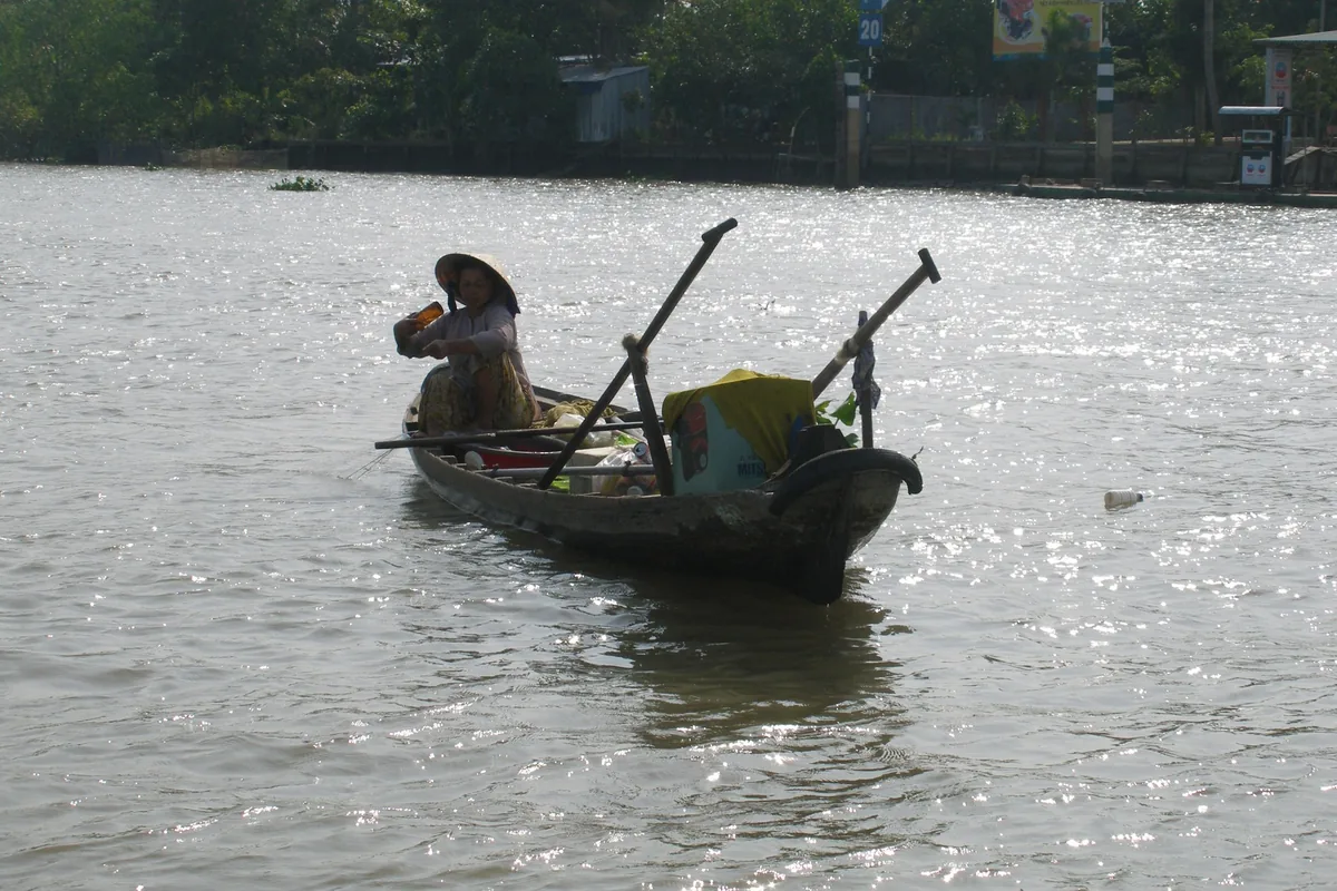 A woman in a conical hat rows a small wooden boat loaded with goods on the Mekong Delta river.
