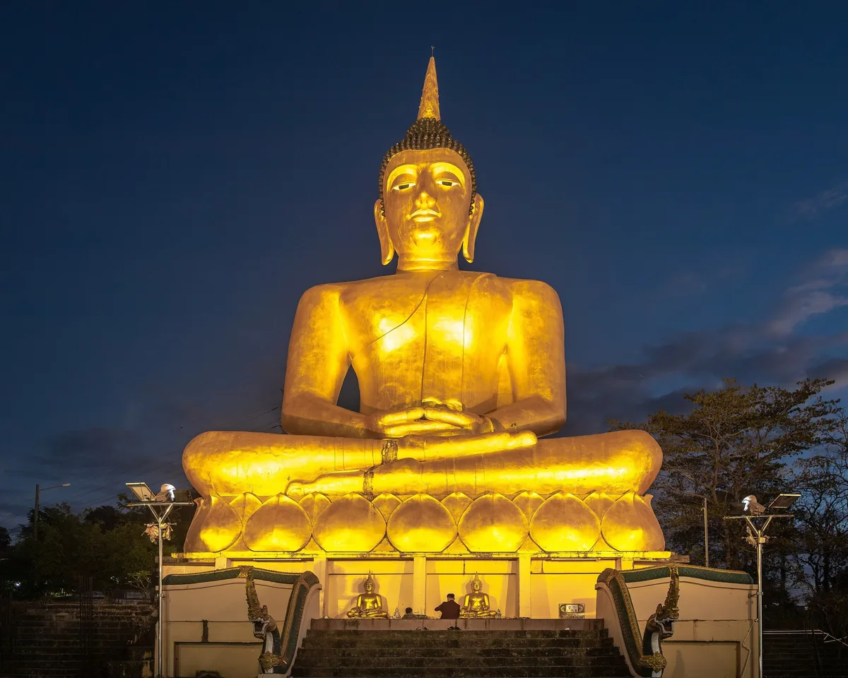 A giant golden seated Buddha statue glows against the dusk sky atop Wat Phou Salao in Pakse.
