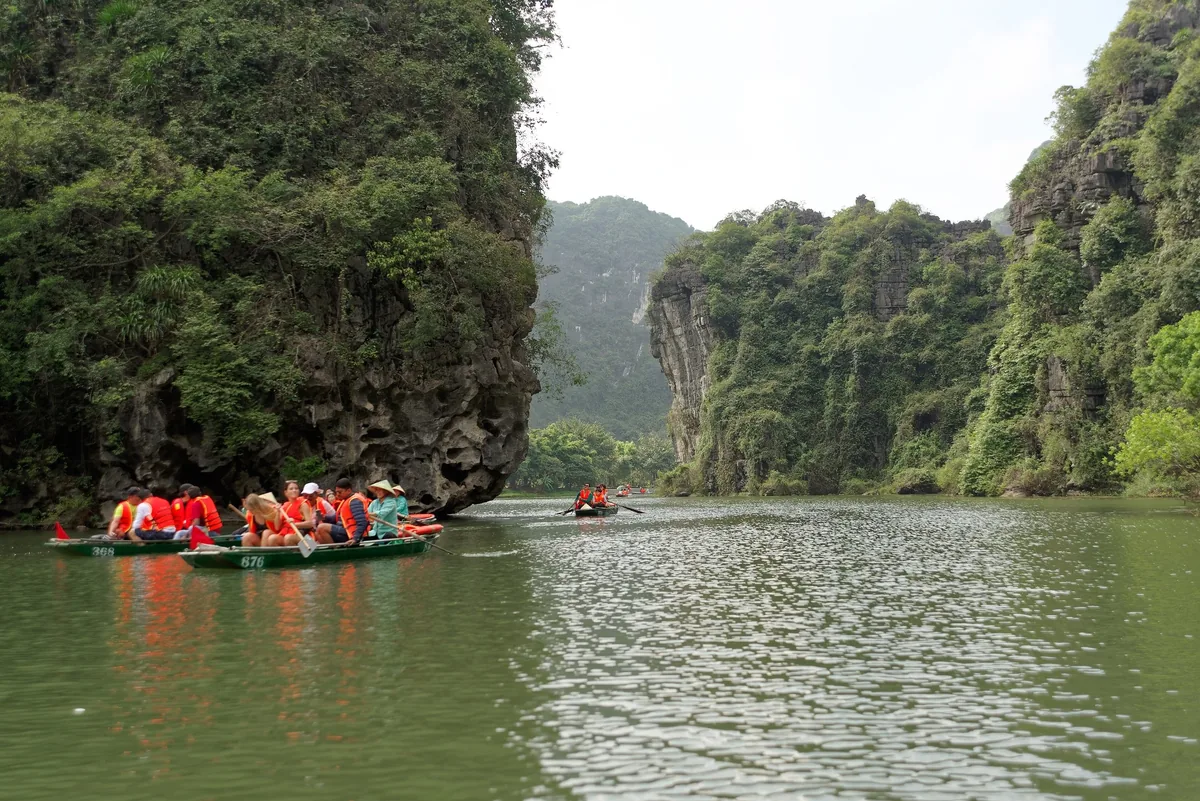 Tourists in orange life jackets row small boats through limestone karst cliffs on the river at Trang An, Ninh Binh.