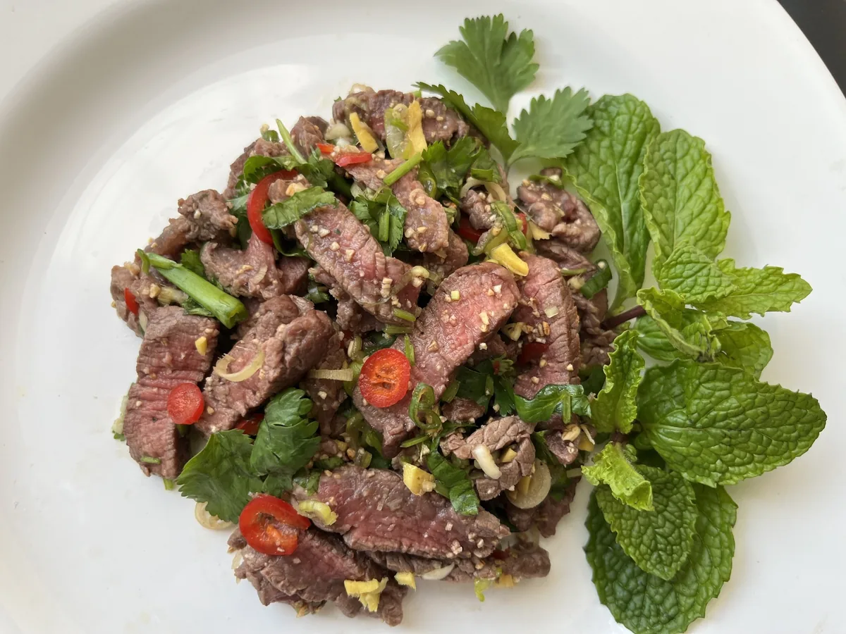 Sliced grilled beef nam tok salad with chili, herbs and mint leaves on a white plate in Laos.