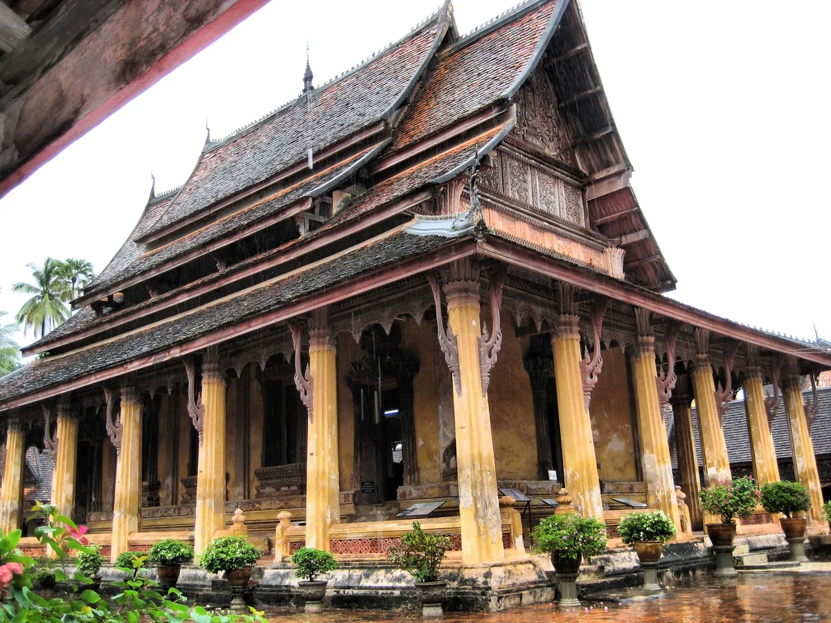 A traditional Lao Buddhist temple with steep tiered roofs and golden columns stands in Vientiane on a rainy day.