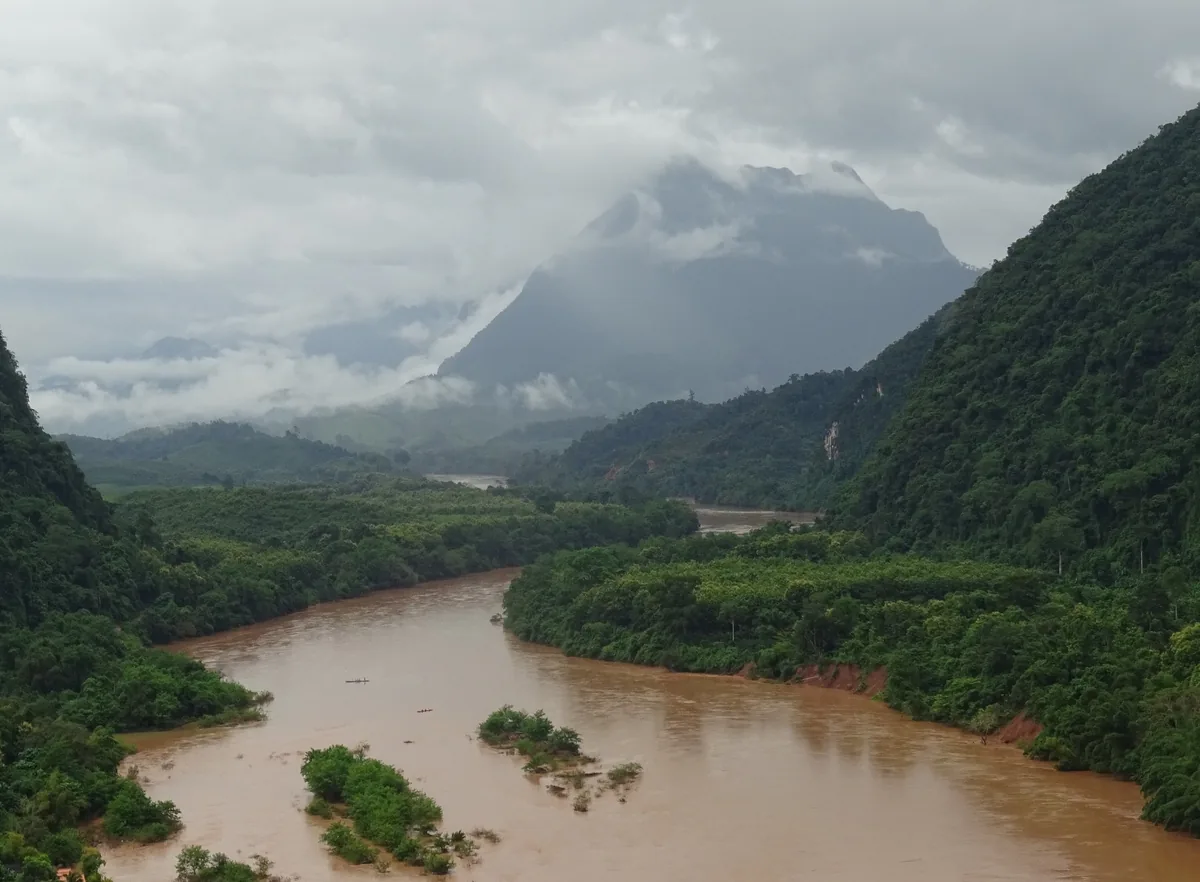 A muddy river winds through jungle-covered mountains with a mist-shrouded peak in Nong Khiaw, Laos.