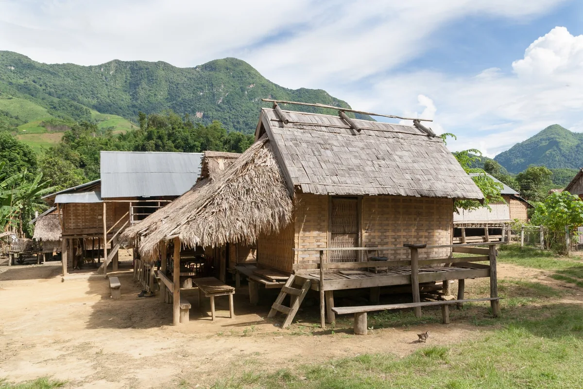 Traditional bamboo stilt houses with thatched roofs in a village near Nong Khiaw, Laos, backed by green mountains.