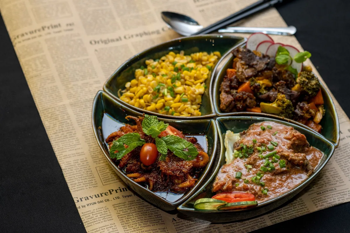A four-section platter of Myanmar dishes with corn, stir-fried beef, crispy salad, and curry served on newsprint paper