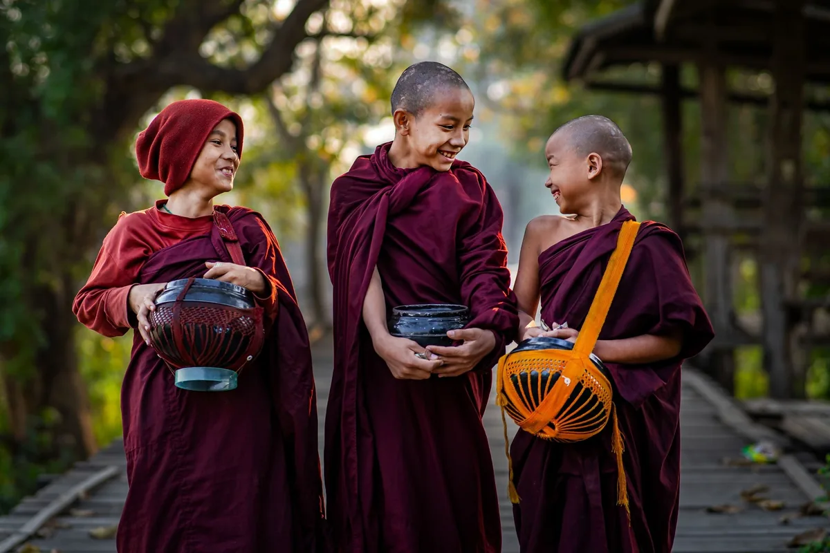 Three young Buddhist monks in maroon robes share a smile while carrying alms bowls on a wooden bridge in Myanmar.