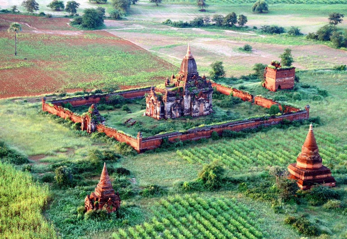 An aerial view shows a walled brick temple compound surrounded by green fields and scattered stupas near Bagan.