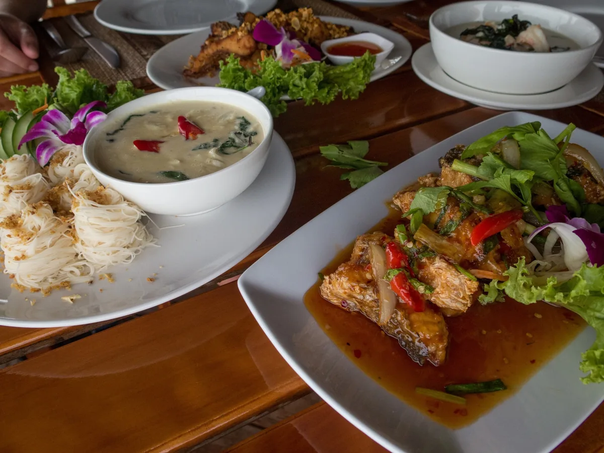 A Thai meal spread on a wooden table features coconut soup, rice noodle rolls, and spicy fried fish with fresh garnishes.