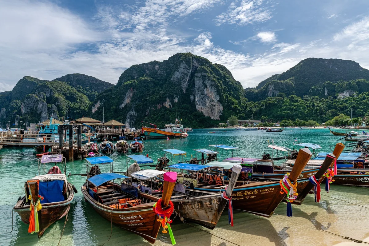 Colorful longtail boats moored in turquoise water beneath dramatic limestone cliffs in Thailand.