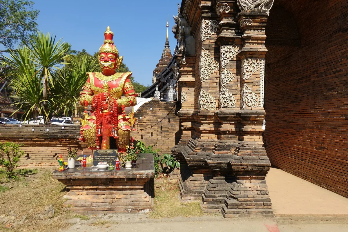 A red and gold guardian statue stands beside an ornate brick chedi at a temple in Chiang Mai.